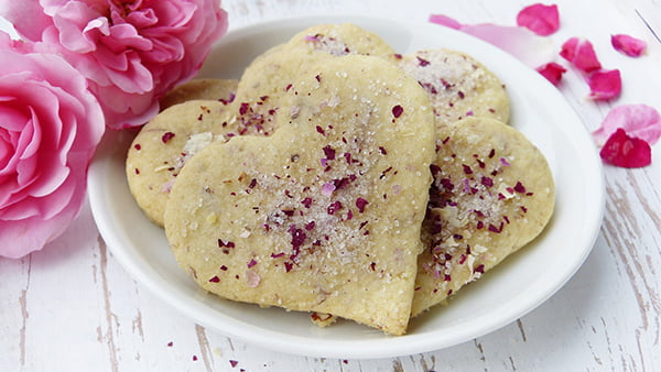 Herzkekse mit rosenblättern und zucker bestreut auf einem kleinen weißen teller mit rosen als deko drumherum