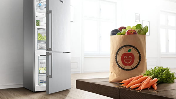 papiertüte mit apfelgesicht und möhren davor mit silbernem kühlschrank im hintergrund