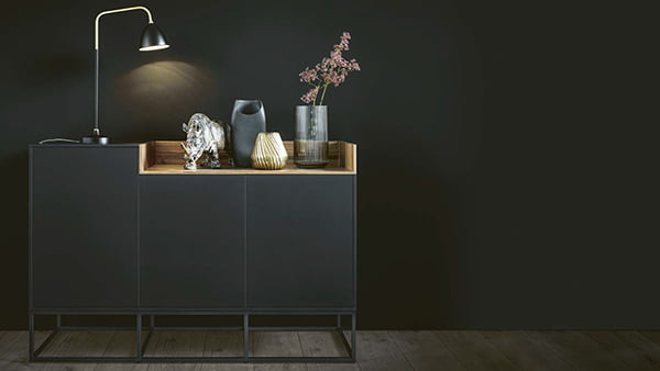 dunkles next125 sideboard mit holzeinsatz blume und tischlampe vor schwarzer wand mit highlight beleuchtung