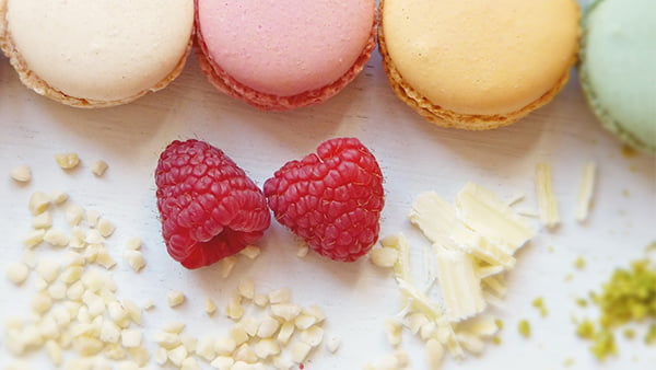 zwei Erdbeeren und Mandeln vor einer Reihe verschieden farbiger Macarons
