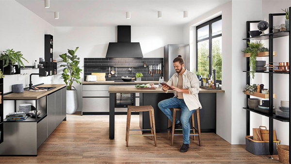 mann sitzt auf einem holzhocker an der nobilia kochinsel in einer industrial style küche mit stahl küchenfront